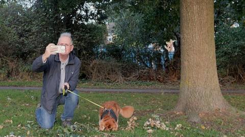 A man in a blue coat and blue jeans with a small brown dog next to a tree taking a photograph