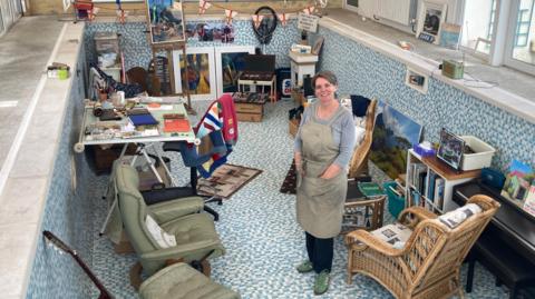 A woman with short grayish brown hair standing in a pool which is filled with various chairs, equipment and paintings. It's arranged so it creates an art studio.