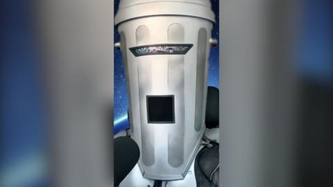 Count Binface wears a helmet that looks like a metal waste bin with grilles for his eyes and mouth, and speaks into a mic.