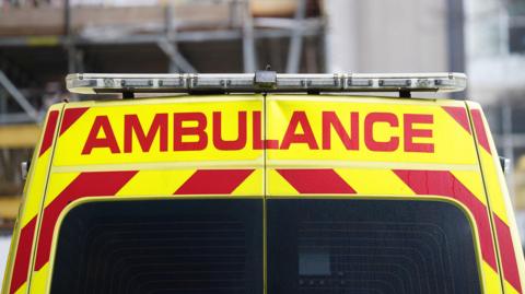 Photo shows the rear of a UK ambulance. The vehicle is yellow and red with "ambulance" written in capital red lettering. 