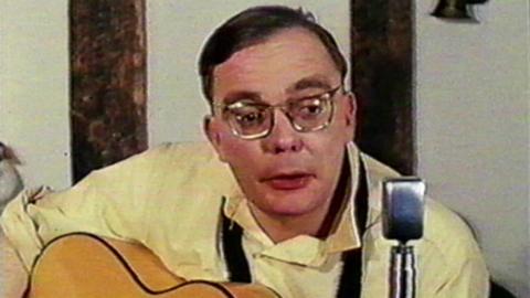 A man wearing a cream shirt, spectacles, and with an acoustic guitar on his lap, sings into an old-fashioned silver microphone. He has dark hair swept to one side.