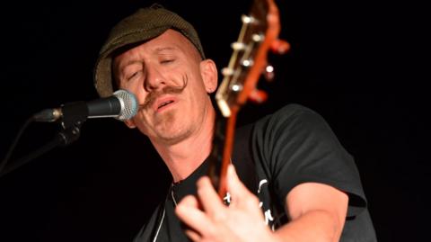 Foy Vance has a handlebar moustache and is wearing a tweed flat cap and a black short-sleeve t-shirt. He is holding a guitar out in front of him and his left hand is on the fretboard. To the right of his mouth is a microphone that he is singing into. 