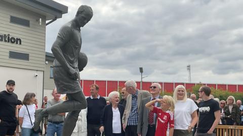 A dark-coloured statue of a footballer, in movement, holding a football. There is a crowd gathered round it on a grey day