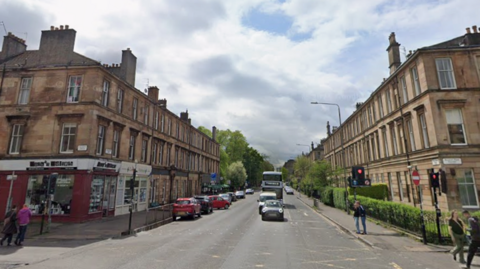 A wide street in south Glasgow lined with sandstone tenement buildings on both sides, with shops at street level. Cars and a bus travel along the road, while pedestrians wait at traffic lights and walk along the pavements. Trees and hedges add greenery near the junction under a partly cloudy sky.