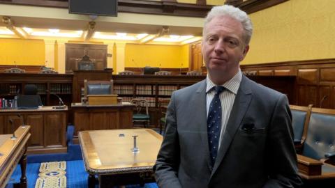 Alex Allinson, who is wearing a grey suit, striped shirt and blue spotted tie and has grey hair, standing in the Tynwald chamber, which has a large brown desk, blue carpet and blue leather chairs.