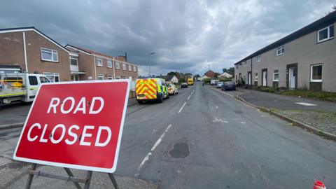 A police sign reading 'road closed' in front of a residential street. Police vans can be seen along with cars.