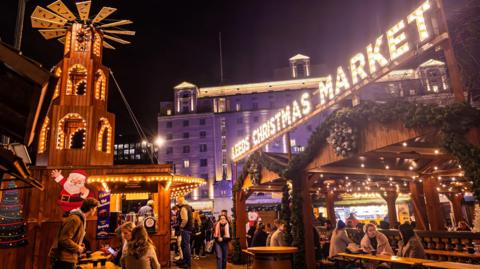 Wooden chalets in Leeds city centre. The chalets are decorated with lights and Christmas garlands. There is a sign lit up which says 'Leeds Christmas Market' along with crowds of people who are either stood up or sat down dressed in winter clothing. 