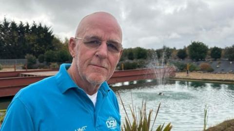 Mike Richardson, who has white stubble and is wearing glasses and a bright blue polo shirt saying DSM in white letters. He is standing outside in front of a pond, with a fountain, behind his aircraft hangar.