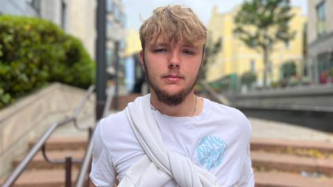 Boy with a short hair but a long blonde fringe and beard looking at the camera. He is wearing a white t-shirt with a blue logo on the right and a white jumper tied over his shoulder.
