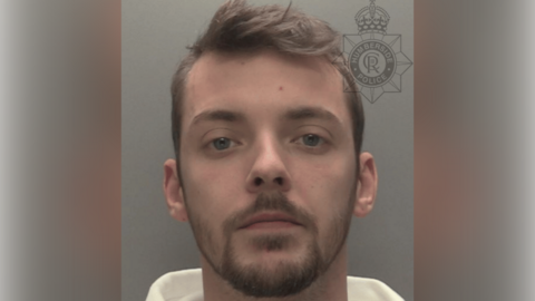 A police custody shot of Jacob Agnew. He is wearing a white hoodie and has short brown hair and short brown facial hair. He is looking at the camera. The wall behind him is grey.