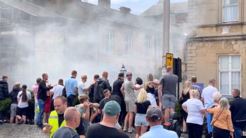 Groups of people throwing bottles and other items at a hotel with smoke in the background