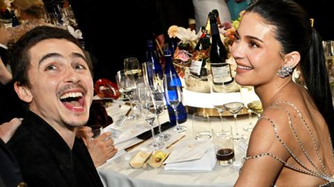Timothee Chalamet and Kylier Jenner are seated at what looks like a formal dinner or gala. Chalamet on the left is laughing openly, leaning slightly forward with clear enjoyment. Jenner beside him is smiling, dressed in an elegant gown with detailed beaded straps and large decorative earrings. The table in front of them is set with multiple wine glasses, bottles of water and champagne, floral arrangements, and plates of food. The overall atmosphere suggests a celebratory or high‑profile event, with warm lighting and a sense of lively conversation around them.