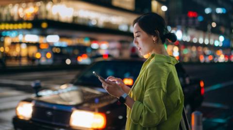 A woman with dark hair wearing a green shirt waits at the side of the road for a taxi at night. Behind her, out of focus, is a street full of bars.