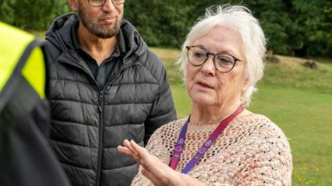 Danielle Stone, to the right, with her hand out, wearing a knitted top, with a lanyard round her neck. She has on glasses and has short fair hair. She is standing by a bearded man wearing a black coat, a black T-shirt, wearing glasses. He is looking at her. They are in a field with trees and grass behind them. 