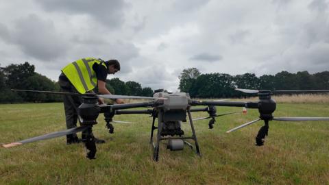 A man wearing a yellow fluorescent safety jacket leans over a large drone to fold out a rotor arm