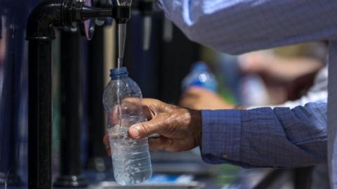 The arms of a man wearing a shirt can be seen filling up a water bottle. 