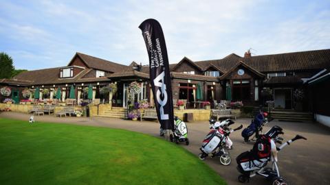 A single storey clubhouse with steps and tables leading to a path and green grass to the left. There are three golf bags on trolleys on the pathway