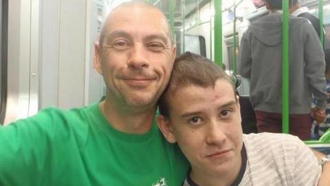 Colin and Rhys seated together on an underground train. Colin wears a bright green t-shirt bearing a Fox logo, and smiles at the camera. He has one arm around his son's shoulder and one arm on the carriage window sill. Rhys is wearing white and grey stripped t-shirt and has his head on his father's shoulder. 