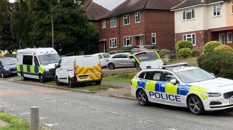 Two police cars and a white van parked on verges on a street with houses behind them