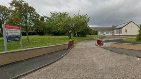 A Google Streetview screenshot of the former St Mary's Church of England Middle School in Belford, Northumberland. A concrete driveway leads to an L-shaped cream building with large windows. There is a sign with the name of the school at the side of the road.