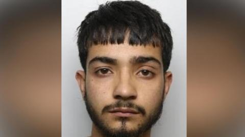 A man with a short black beard and goatee. He has short straight black hair cut straight across is forehead. He has an unhappy expression.