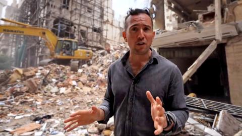 Middle East correspondent Hugo Bachega gestures while speaking to camera in front of a digger and rubble on a residential street in Beirut.