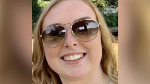 A photograph of Stephanie Irons, a 23-year-old white woman. Se is smiling, wearing aviator style sunglasses and has wavy hair