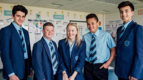 Five high school pupils stand in a classroom smiling at the camera - a mix of boys and girls.