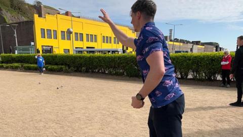 A man wearing a navy blue shirt with pink fish on it throws a petanque ball during a game. A building with bright yellow walls is in the background.