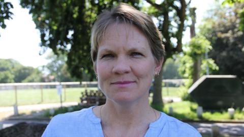 A picture of Deputy Sasha Kazantseva-Miller. She has short brown hair and is standing in front of a large field.