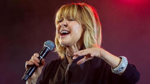 Lulu has her eyes shut as she belts out one of her songs into a microphone.