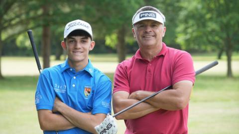 Two Norfolk golfers stand side-by-side at Dereham Golf Club. Teenager Liam Caffyn on the left in a blue golf shirt with a Norfolk crest and professional Andrew Marshall wearing a red shirt. Both are smiling at the camera. They are standing in front of a tree-lined fairway. 
