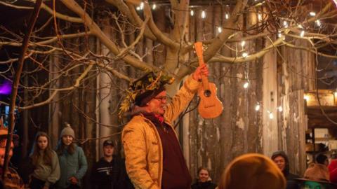 A man wearing a top hat with a wreath of twigs on its brim is holding a ukulele. There are people around him and two fire torches are raised.