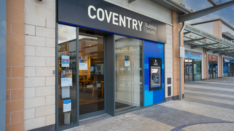 A branch of Coventry Building Society, with those words on the outside in white on a black background above the front door. A cash machine outside is to the right of the door and different outlets are on the far right of the photo. 