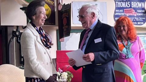 Princess Anne smiling with Malcolm Stern who is also holding and holding a certificate. They are on stage together at an event.