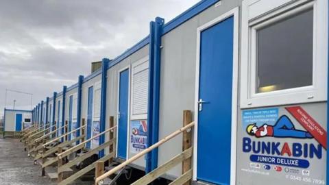 External view of the row of ten pods, grey portable cabins with wooden steps up to a blue front door