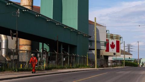 An image of a steel plant in Hamilton, Ontario. A sole worker in a red jumpsuit and a helmet is seen walking outside. The plant building is green, white and beige, and features a large Canadian flag on one panel.