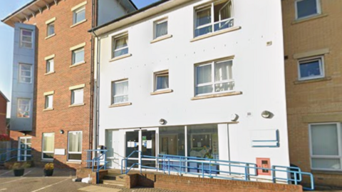 The Family Hub in Penrith is a three-storey white block between two larger, red brick buildings. There is a ramp and steps leading to the front entrance which has automatic doors and large windows.. 