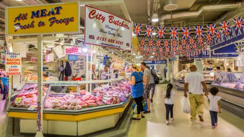 The inside of Birmingham Indoor Market with a meat stand and people and union jack flags. 