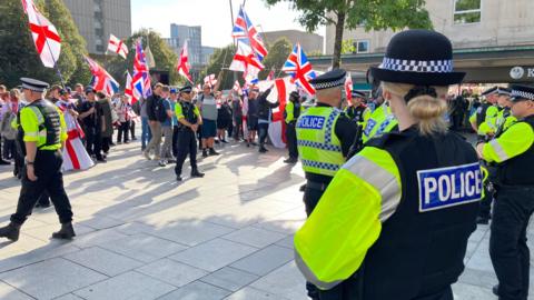 Two rows of uniformed police officers are standing watching a group of about 50, mostly men, many of whom are holding St George and Union Jack flags in a street in Plymouth. 