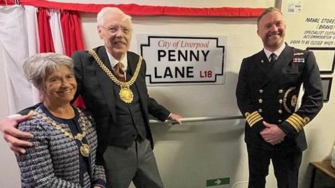 Richard and Erica Kemp, wearing gold mayoral chains, stands next to the Penny Lane street sign, which has been unveiled behind a red and white curtain. Capt Will Blackett, in his naval uniform, smiles at the camera 