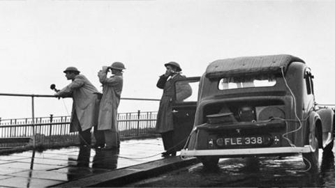 Man holding microphone looks out at sea and stands beside two other men and an old fashioned car.