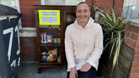 Sarah Dolan with brown hair tied back, wearing a beige hoodie. She is kneeling down, smiling, next to a brown glass cabinet full of food items. It is in front of her house with a sign on it stating "Avenues & Dukeries Community Pantry".  