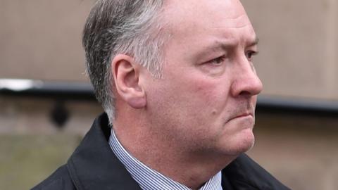 A profile close-up of Ian Paterson who has short, grey hair and is wearing a striped shirt and a black jacket.