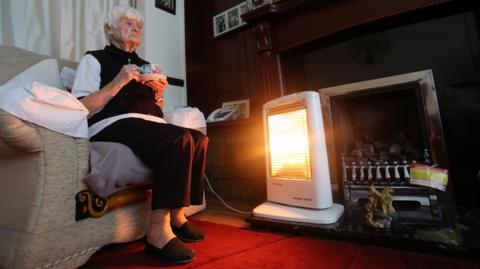 An elderly lady sitting on a sofa in a lounge holding a cup of tea in front of an electric fire