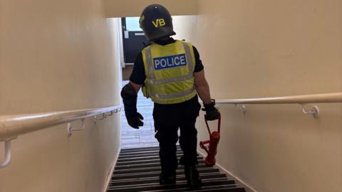 The back of a police officer with a high-vis vest on walking down the stairs. He is holding a door ramming device. The stairs have two rails on either side and the walls are cream. A door is at the bottom. 