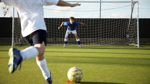 Kid kicking a football at a goalkeeper in a goal