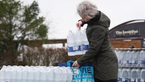 A woman collects bottled water from a water station in East Grinstead, she is wearing a dark coat 