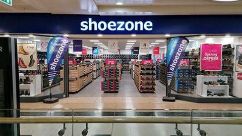 A shoe zone store inside a shopping centre. Stacks of shoes can be seen through a shop window with the shoe zone signs across the window and outside the store