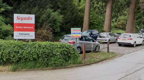 The entrance to the Kirkby Malzeard creamery plant. The picture shows the sign reading "Saputo. Kirkby Malzeard. Entrance. Visitors please report to reception". The picture also shows a health and safety sign and several parked cars. 